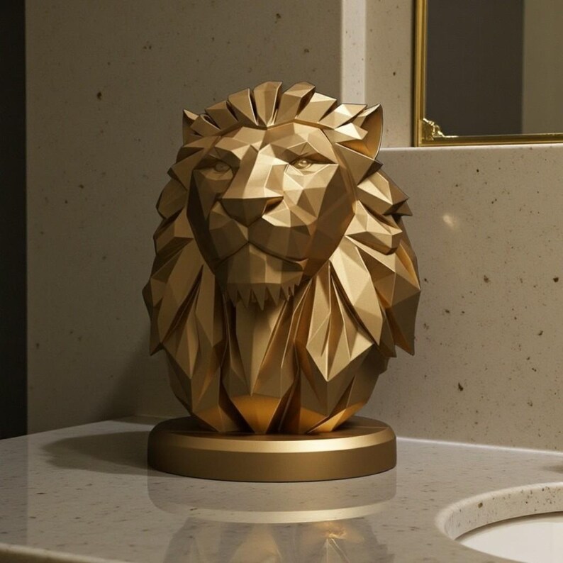 May include: A gold-colored, geometric lion head sculpture. The sculpture is made of many triangular facets, giving it a modern, angular appearance. It sits on a round base, and the overall design is symmetrical and detailed. The lion's mane and facial features are clearly defined.