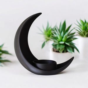 Puede incluir: Un portavelas negro en forma de media luna. Tiene una hendidura circular para sostener una vela. El fondo presenta varias plantas verdes peque&ntilde;as en macetas. Un art&iacute;culo decorativo.