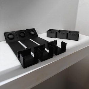 Może przedstawiać: Cztery czarne plastikowe wsporniki półkowe z hakiem na końcu. Wsporniki są zaprojektowane do trzymania przedmiotów na półce.