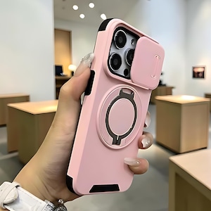 May include: A light pink smartphone case featuring a sliding camera lens cover and a circular magnetic ring stand on the back. The case has black accents along the edges and corners, providing a contrast to the matte pink finish of the protective cover.