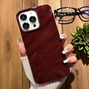 K&ouml;nnte beinhalten: Eine weinrote Handyh&uuml;lle mit gl&auml;nzendem Finish wird in der Hand gehalten. Die H&uuml;lle hat einen Kameraausschnitt mit drei Linsen und einem Blitz. Eine schwarze Brille und eine gr&uuml;ne Sukkulente sind im Hintergrund.