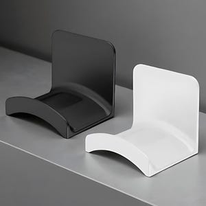 May include: Two modern, minimalist headphone stands. One is glossy black, the other is glossy white. Each stand has a curved base and a vertical back, designed to hold headphones. The stands are displayed on a gray surface.