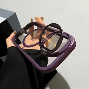 Peut inclure: Un étui de téléphone violet avec un support pliable intégré et un protecteur d'objectif transparent. Les mots "CASE PRO" sont imprimés sur le support. L'étui est maintenu, montrant le support en position ouverte.