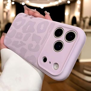 May include: A lavender phone case with a leopard print design. The case has a smooth finish and features three camera cutouts and a flash cutout. The case is held in a hand.