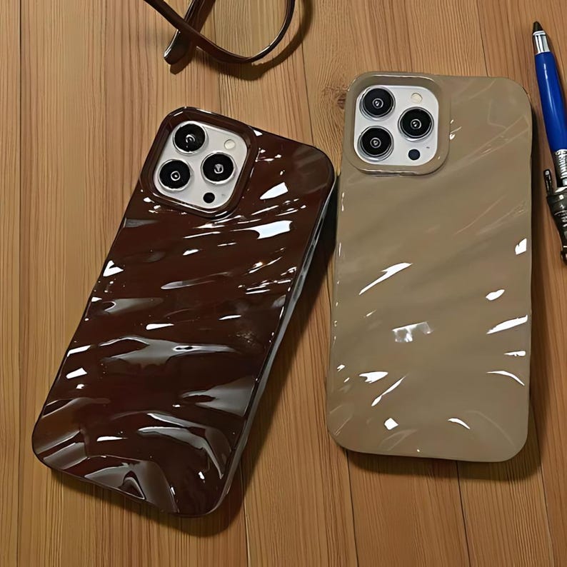 K&ouml;nnte beinhalten: Zwei gl&auml;nzende Handyh&uuml;llen. Eine ist dunkel schokoladenbraun, die andere hellbeige. Beide haben ein wellenf&ouml;rmiges, strukturiertes Design und liegen auf einer Holzoberfl&auml;che. Die H&uuml;llen haben Kameraausschnitte.