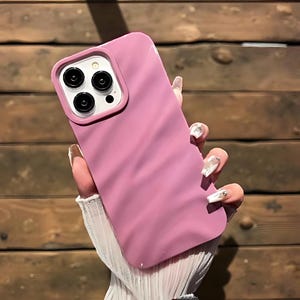 K&ouml;nnte beinhalten: Eine rosa Handyh&uuml;lle mit welliger Textur wird in einer Hand gehalten. Die H&uuml;lle hat einen Kameraausschnitt mit drei schwarzen Linsen und einem wei&szlig;en Kreis. Der Hintergrund ist eine Holzoberfl&auml;che.