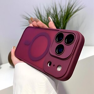 Puede incluir: Una funda de tel&eacute;fono burdeos con un &aacute;rea de carga MagSafe circular transparente. La funda tiene una secci&oacute;n elevada para la c&aacute;mara con tres lentes y un flash. La funda se sostiene en una mano, con un fondo borroso de hojas verdes.