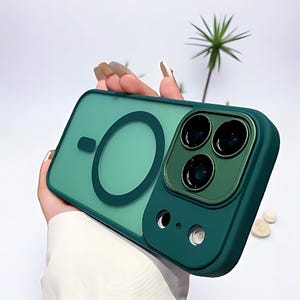 Puede incluir: Una funda de tel&eacute;fono verde azulado con un anillo magn&eacute;tico y protector de lente de c&aacute;mara. La funda tiene una parte trasera transl&uacute;cida y un borde verde oscuro. El protector de lente tiene tres aberturas circulares.