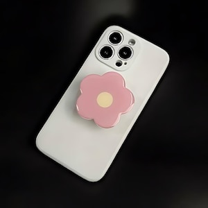 May include: A white smartphone with a pink flower-shaped phone grip. The phone has a triple-lens camera and a glossy finish. The flower grip has a light yellow center.