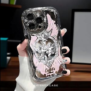 May include: A silver phone case with a pink backdrop, showcasing a detailed cherub illustration. The case has a decorative border and the words "Cupid's Angel" are visible. The brand name "WHISKERCASE" is printed at the bottom.
