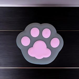 Könnte beinhalten: Eine graue, pfotenförmige Matte mit rosa Pfotenballen. Die Matte besteht aus einem weichen Material und dient zum Schutz von Oberflächen. Das Pfotenabdruck-Design ist eine niedliche und verspielte Ergänzung für jeden Raum.