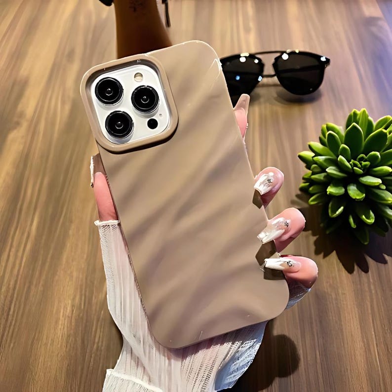 K&ouml;nnte beinhalten: Eine beige Handyh&uuml;lle mit einem Wirbeldesign. Die H&uuml;lle hat einen Kameraausschnitt und einen sch&uuml;tzenden Rand. Sonnenbrille und eine Sukkulentenpflanze befinden sich im Hintergrund.