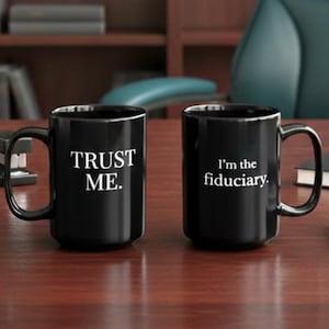 May include: Two black ceramic mugs with white text on a wooden desk. One mug says "TRUST ME." and the other says "I'm the fiduciary." The mugs have a glossy finish and are in a professional setting.