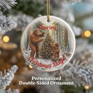 May include: A round white ceramic ornament featuring a printed illustration of two brown Sasquatch figures decorating a Christmas tree in a snowy forest. The red text "Sharing Christmas Magic" is printed on the front. It hangs from a gold string.