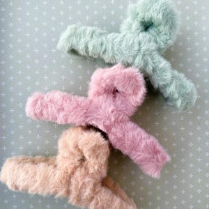 Könnte beinhalten: Drei flauschige Haarklammern in verschiedenen Farben: hellgrün, rosa und beige. Die Klammern sind knotenförmig und aus weichem, flauschigem Material gefertigt.
