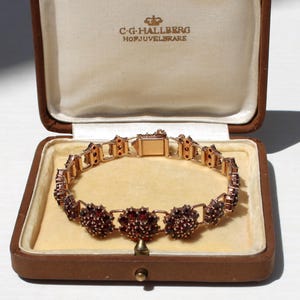 Pode incluir: Uma pulseira dourada com pedras preciosas vermelhas, exibida numa caixa de joias castanha com interior creme. A pulseira apresenta um design floral e um fecho. A caixa está aberta, revelando a pulseira.
