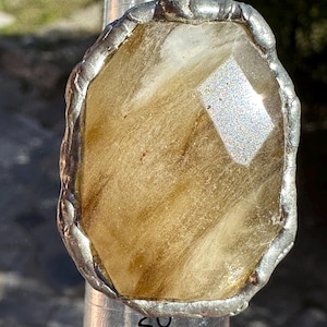 May include: A handcrafted ring featuring a large, faceted, oval-shaped gemstone with a warm, amber hue and natural inclusions. The stone is set in a silver-colored, irregular bezel. The ring is shown on a ring sizer.