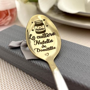 Könnte beinhalten: Ein goldfarbener Löffel mit weißem Griff, graviert mit den Worten "La cuillère Nutella de Domitille" und einer Nutella-Glas-Illustration. Der Löffel wird in einer grauen Geschenkbox mit einem grauen Band präsentiert.