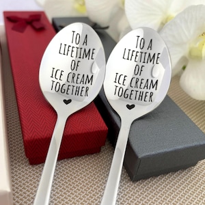 May include: Two stainless steel spoons with the text "TO A LIFETIME OF ICE CREAM TOGETHER" and a heart symbol. The spoons are displayed in front of red and gray gift boxes, suggesting a gift item.
