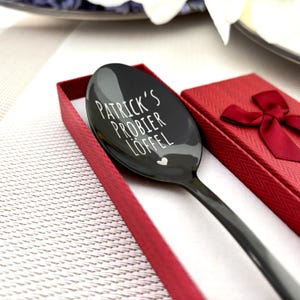 Può includere: Un cucchiaio nero con la scritta bianca "PATRICK'S PROBIER LÖFFEL", presentato in una confezione regalo rossa. Un piccolo cuore bianco è inciso sotto il testo. Il cucchiaio è in una scatola rossa con un nastro abbinato.