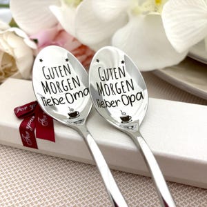 Peut inclure: Deux cuillères en argent gravées avec "Guten Morgen liebe Oma" et "Guten Morgen lieber Opa" en noir. Chaque cuillère présente un motif de tasse à café et des étoiles. Les cuillères sont présentées dans un coffret cadeau blanc avec un ruban rouge.