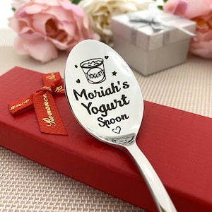 May include: A stainless steel spoon engraved with "Moriah's Yogurt Spoon" and a yogurt cup graphic. The spoon is on a red gift box with a "Romance" ribbon, with pink and white flowers and a gift box in the background.