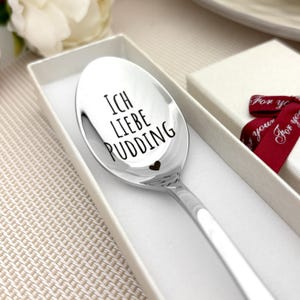 Könnte beinhalten: Ein glänzender, silberner Löffel mit der Gravur "ICH LIEBE PUDDING" und einem kleinen Herz. Der Löffel liegt in einer weißen Geschenkbox mit rotem Band.