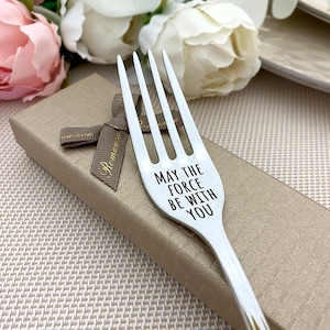 Può includere: Una forchetta d'argento a quattro rebbi, incisa con la frase "MAY THE FORCE BE WITH YOU" in nero. La forchetta è appoggiata su una confezione regalo dorata con un nastro marrone. Decorazioni floreali sullo sfondo.