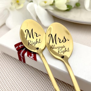 May include: Two gold-coloured spoons, each inscribed with black script: "Mr. Right" and "Mrs. Always Right". Small heart details are present on each spoon. They rest on a white box, accented by a red ribbon.
