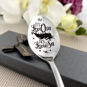Può includere: Un cucchiaio d'argento con la frase "It's Leviosa Not Leviosa" incisa, insieme a un design di piuma e stelle. Il cucchiaio è presentato in una confezione regalo con un nastro.