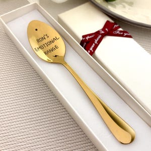 May include: A gold-colored spoon with the text "Ron's Emotional Range" engraved on the spoon's head. The spoon is inside a white box with a red ribbon tied around it. A star and lightning bolt are also engraved on the spoon.