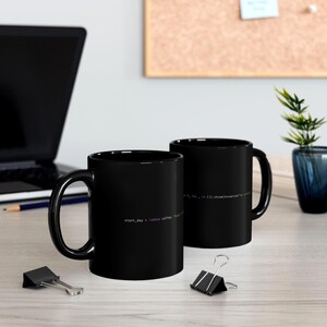May include: Two black coffee mugs with white text featuring code snippets. One mug reads "start_day = lambda coffee:" in purple and green. The other mug has code in white. The mugs are on a light-colored surface.