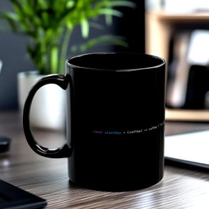 May include: A black ceramic coffee mug with a curved handle. The mug features the text "const startDay = (coffee) => coffee ?" in a colorful, stylized font. The mug is on a wooden surface.