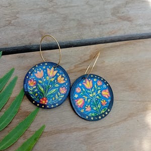Könnte beinhalten: Ein Paar runde, dunkelblaue Holzohrringe mit handgemalten Blumenmotiven aus orangefarbenen, gelben und grünen Tulpen und Blättern. Die kreisförmigen Scheiben sind mit kleinen gelben Punkten an den Rändern verziert und hängen an dünnen, goldfarbenen Metallreifen.