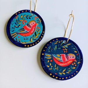 Puede incluir: Un par de pendientes de aro circulares con diseños pintados a mano de pájaros rojos rodeados de follaje verde y pequeñas bayas naranjas sobre un fondo azul verdoso, enmarcados por un borde azul oscuro con puntos dorados y aros de metal.