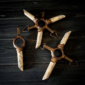 Puede incluir: Tres objetos decorativos en forma de estrella de color bronce con detalles texturizados en blanco roto. Cada pieza tiene un centro circular y un asa. Los objetos están dispuestos sobre una superficie de madera oscura.