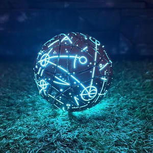 May include: A spherical object with glowing, intricate patterns. The sphere, resting on artificial grass, emits a soft, blue light, creating a mystical ambiance. The dark background enhances the glowing effect of the sphere.