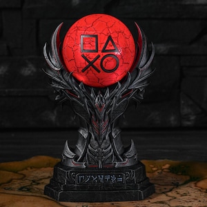 May include: A black and red PlayStation controller symbol sculpture. The sculpture features a cracked red orb with the PlayStation symbols, supported by a detailed, dark-coloured base with a map in the background. The sculpture is approximately 20 cm tall.