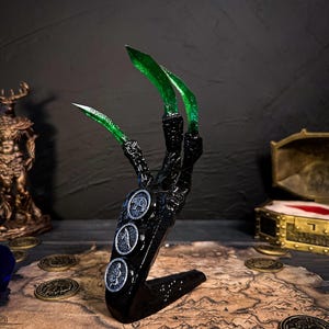 Puede incluir: Una escultura de mano negra con tres garras verdes y puntiagudas. La mano sostiene una base negra con tres emblemas plateados en forma de moneda. La escultura se coloca sobre un mapa con monedas de oro dispersas.