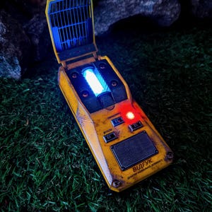 May include: A distressed yellow device with a blue and red light display, featuring a hinged top with a blue grid. The device has a rectangular shape with text and appears to be a prop or model.