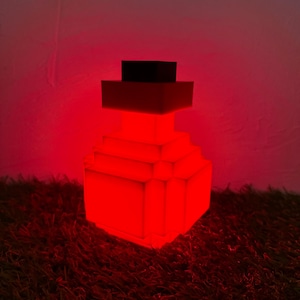 Op de afbeelding: Een rode, gepixelde flesvormige lamp met een zwarte bovenkant. De lamp is verlicht en geeft een rode gloed. De lamp staat op een donkergroen, getextureerd oppervlak.