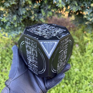 Op de afbeelding: Een zwart, veelzijdig object met ingewikkelde, gegraveerde patronen en een driehoekig ontwerp op één vlak. Het object wordt vastgehouden in een zwarte handschoen, met een groene achtergrond van gebladerte.
