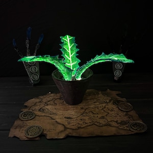 May include: A glowing green plant in a brown pot, set on a map with several bronze coins. The plant has spiky leaves with white veins. The background is dark, with two stylized hand-like objects.