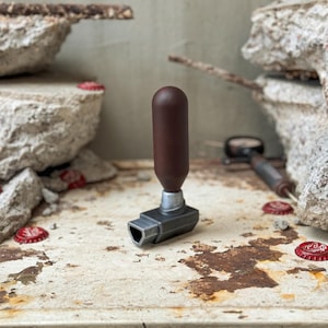 May include: A brown and silver bottle opener with a cylindrical handle. The opener is on a weathered, rusty surface with red bottle caps scattered around. The background features concrete and a neutral wall.