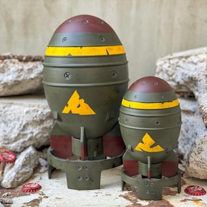 May include: Two olive green, bomb-shaped decorative items with yellow hazard stripes and a yellow triangle symbol. The tops are maroon, and the bases have red accents. The items are on a weathered white surface.