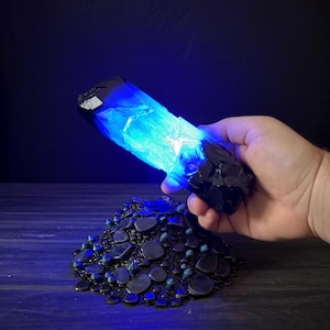 Może przedstawiać: Świecący niebieski kryształ trzymany w dłoni, z ciemną, teksturowaną podstawą z małych, ciemnych kamieni. Kryształ przechodzi od ciemnoniebieskiego do jasnoniebieskiego, emitując miękkie światło. Tło jest ciemnoniebieskie.