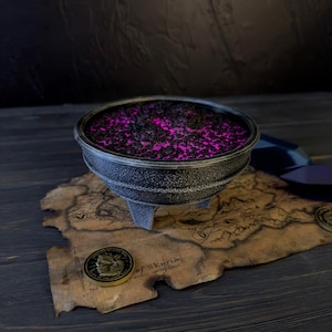 May include: A silver-colored cauldron filled with a vibrant pink substance, resting on a parchment map. The map features a detailed design and text. A gold coin with a detailed relief is also present, suggesting a fantasy or game theme.