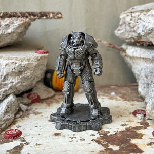 May include: A detailed, metallic grey figurine of a futuristic armoured figure, standing on a textured base. The figure has intricate armour plating and a helmet. The background includes weathered stone and bottle caps, suggesting a post-apocalyptic theme.