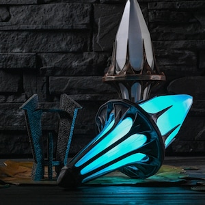 May include: An artistic composition featuring futuristic-looking sculptures. The central piece is a glowing blue, wing-shaped object with black accents. A tall, white and bronze-colored structure stands behind it, and a dark, textured background completes the scene.