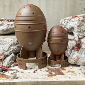 May include: Two brown, bomb-shaped sculptures with a rusted appearance. The larger one is approximately 12 inches tall, and the smaller one is about 8 inches tall. Both have horizontal bands and are mounted on a metal base.